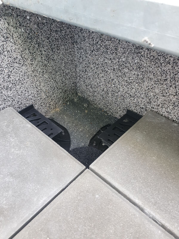 Eckplatzierung zugeschnittener Terrassen-Stelzlager, die angehobene äußere Bodenplatten an einer strukturierten Wand tragen.