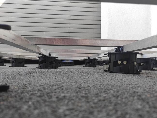 Widok z niskiej perspektywy na aluminiowe legary tarasowe podparte niskoprofilowymi wspornikami regulowanymi na tarasie dachowym.