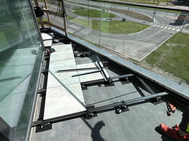 Draufsicht auf die Installation eines erhöhten Balkonbodens mit Porzellanplatten auf Aluminiumbalken und höhenverstellbaren Kunststoff-Stelzlagern.