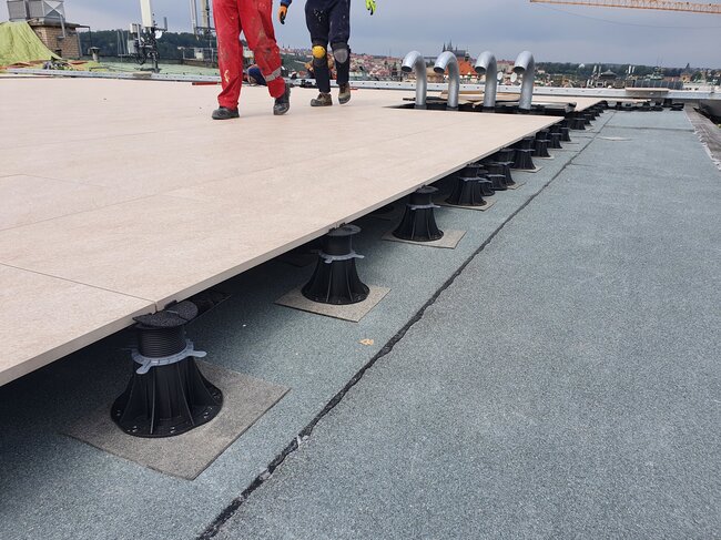 Arbeiter gehen über ein teilweise installiertes System aus erhöhten Terrassenplatten auf einem Dach, das von höhenverstellbaren Stelzlagern getragen wird.