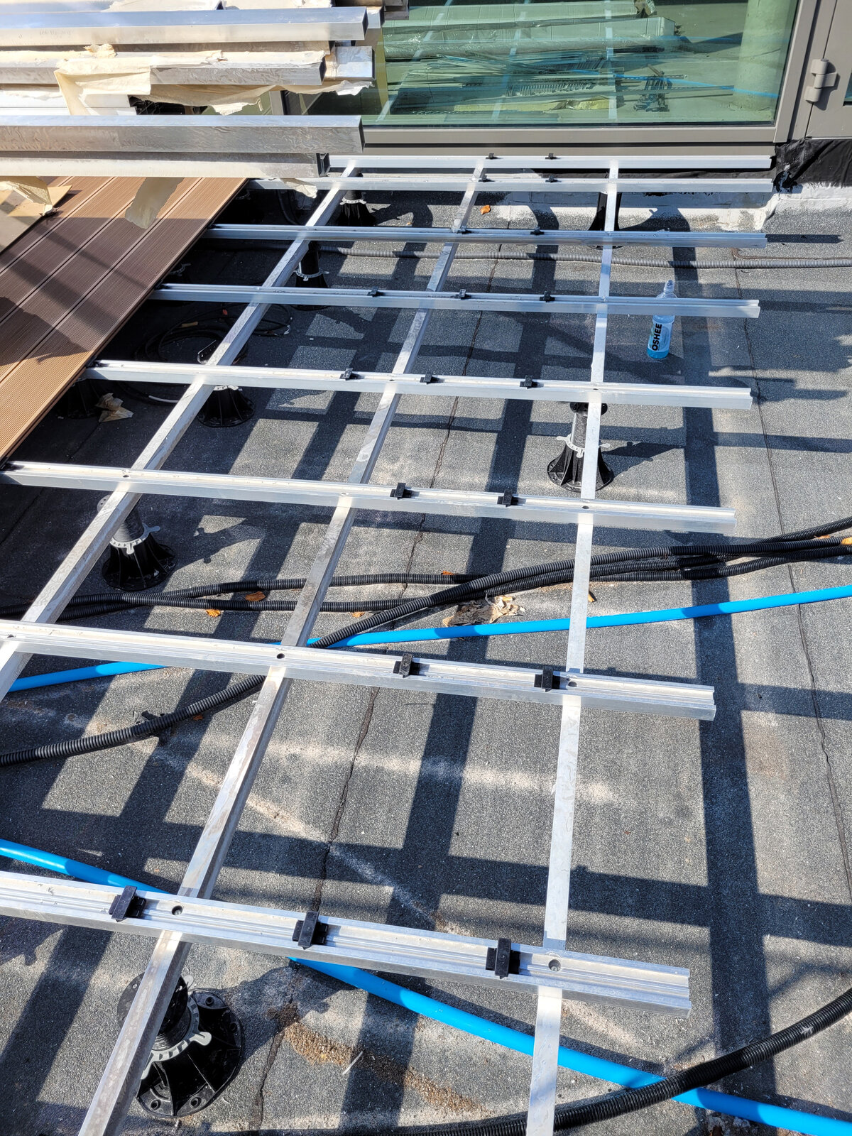 Aluminium-Balkenkonstruktion, montiert auf höhenverstellbaren Stelzlagern, verlegt über Rohren und Verkabelung unter einer erhöhten Terrasse.