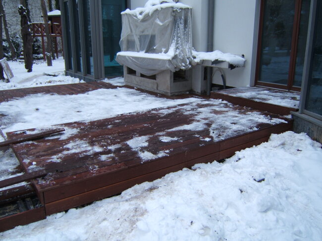 Dunkle Holzterrasse, teilweise mit schmelzendem Schnee bedeckt, vor einem Haus im Winter.