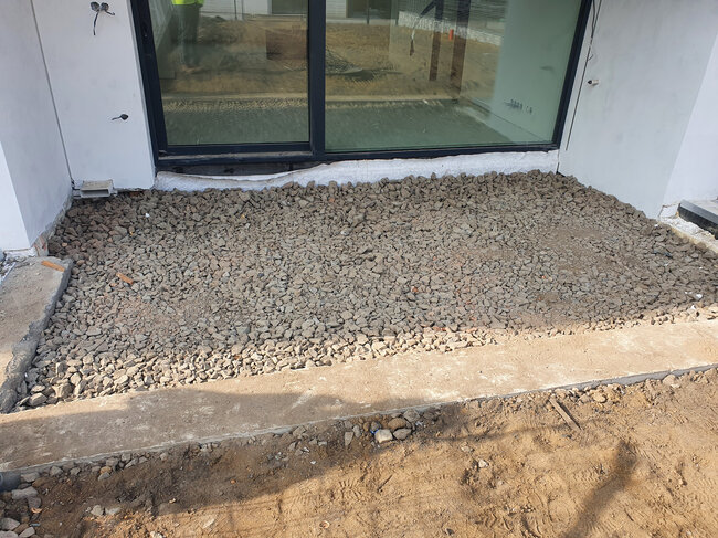 Schicht aus gebrochenem Stein, vorbereitet für den Terrassenbau im Freien vor einer Glasschiebetür.