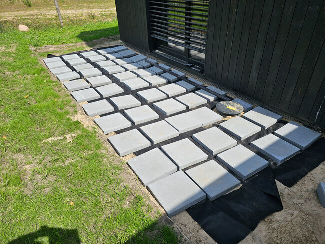 Quadratische Betonplatten, die auf Unkrautvlies verlegt wurden, um den Untergrund einer Außenterrasse vorzubereiten.