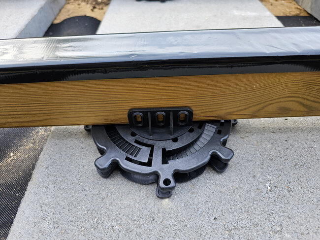 Nahaufnahme eines höhenverstellbaren Kunststoff-Stelzlagers, das einen hölzernen Terrassenbalken trägt.
