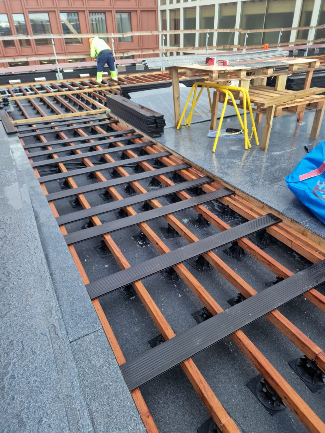 Montage der Unterkonstruktion einer erhöhten Terrasse mit höhenverstellbaren Stelzlagern, die Holzbalken und Verbundwerkstoffdielen tragen.