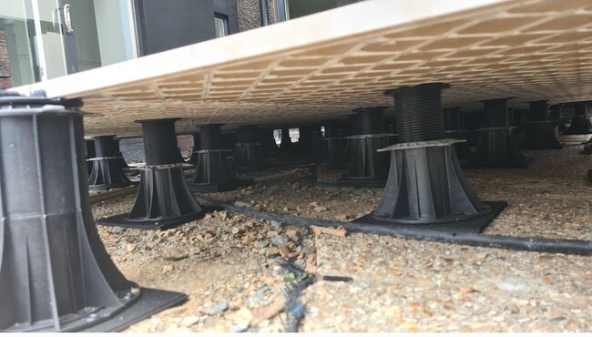 Ansicht von unten auf angehobene Terrassenplatten aus Porzellan, die von höhenverstellbaren Stelzlagern auf Gummipads getragen werden.